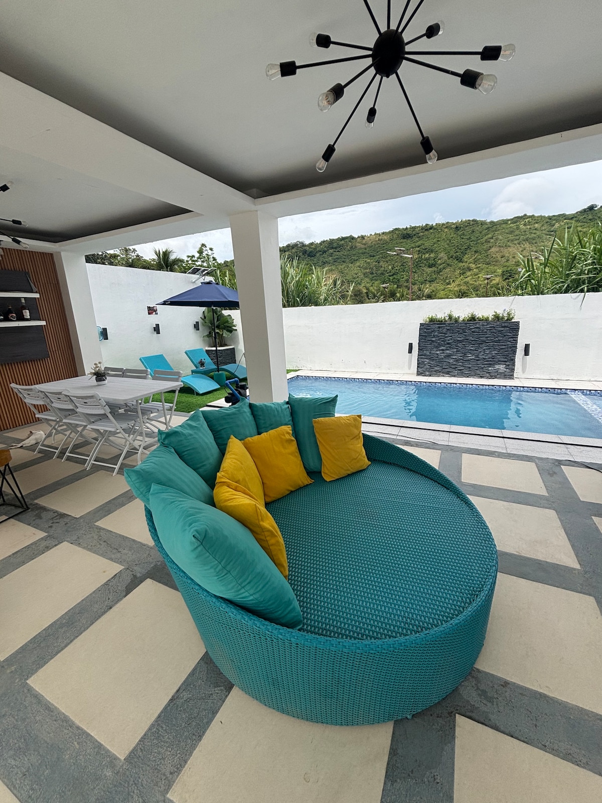 A circular seating area is featured in a vibrant turquoise color, surrounded by bright yellow accent pillows. The seating overlooks a swimming pool, while a backdrop of green hills is visible in the distance. Tables and chairs are arranged nearby, creating an inviting outdoor space.