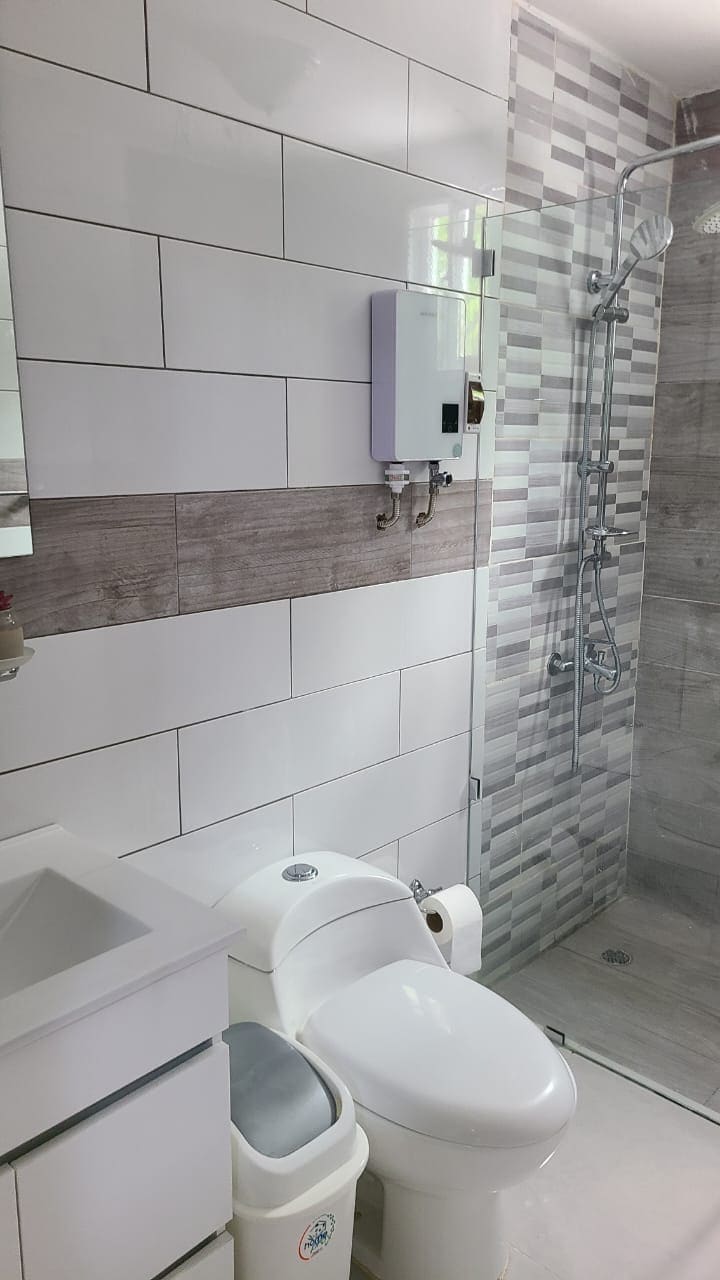 A stylish bathroom features modern tile work with a blend of gray and white tones. A spacious shower with clear glass doors is visible, alongside a sink and toilet. Fresh linens and essential toiletries are provided, creating a clean and functional space.