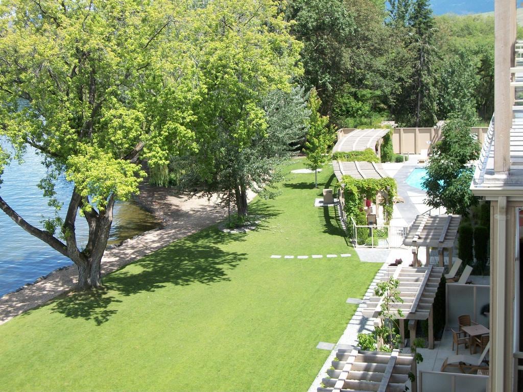 A lush green lawn extends towards a sandy beach by the lake, bordered by trees. A pool is situated nearby, and several pergolas provide shaded seating areas. The serene landscape features a gentle shoreline and a backdrop of trees, enhancing the tranquil outdoor environment.