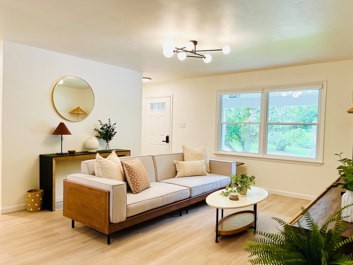A modern living room is presented with a light-colored sofa featuring decorative pillows. A round mirror is mounted above a wooden console table. A small lamp and potted plants add touches of greenery. Large windows allow natural light to illuminate the space.
