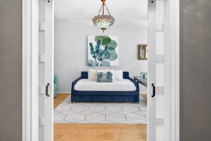 Un dormitorio luminoso y moderno enmarcado por puertas corredizas dobles, con una cama azul intenso, obras de arte botánicas relajantes y un candelabro colorido, que crean un refugio fresco y elegante en el que a los huéspedes les encanta relajarse.