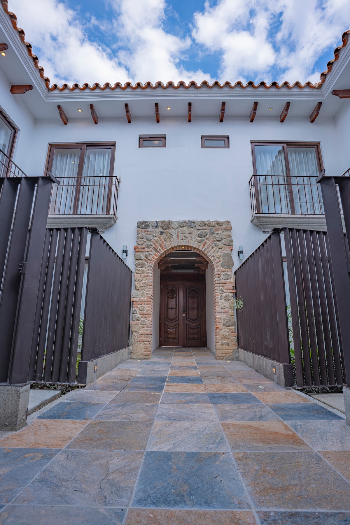 An inviting entrance is framed by stone and wooden elements. The entrance features a large double door with a decorative archway above. Large windows on either side allow natural light to brighten the area, while a textured stone path leads to the doorway.