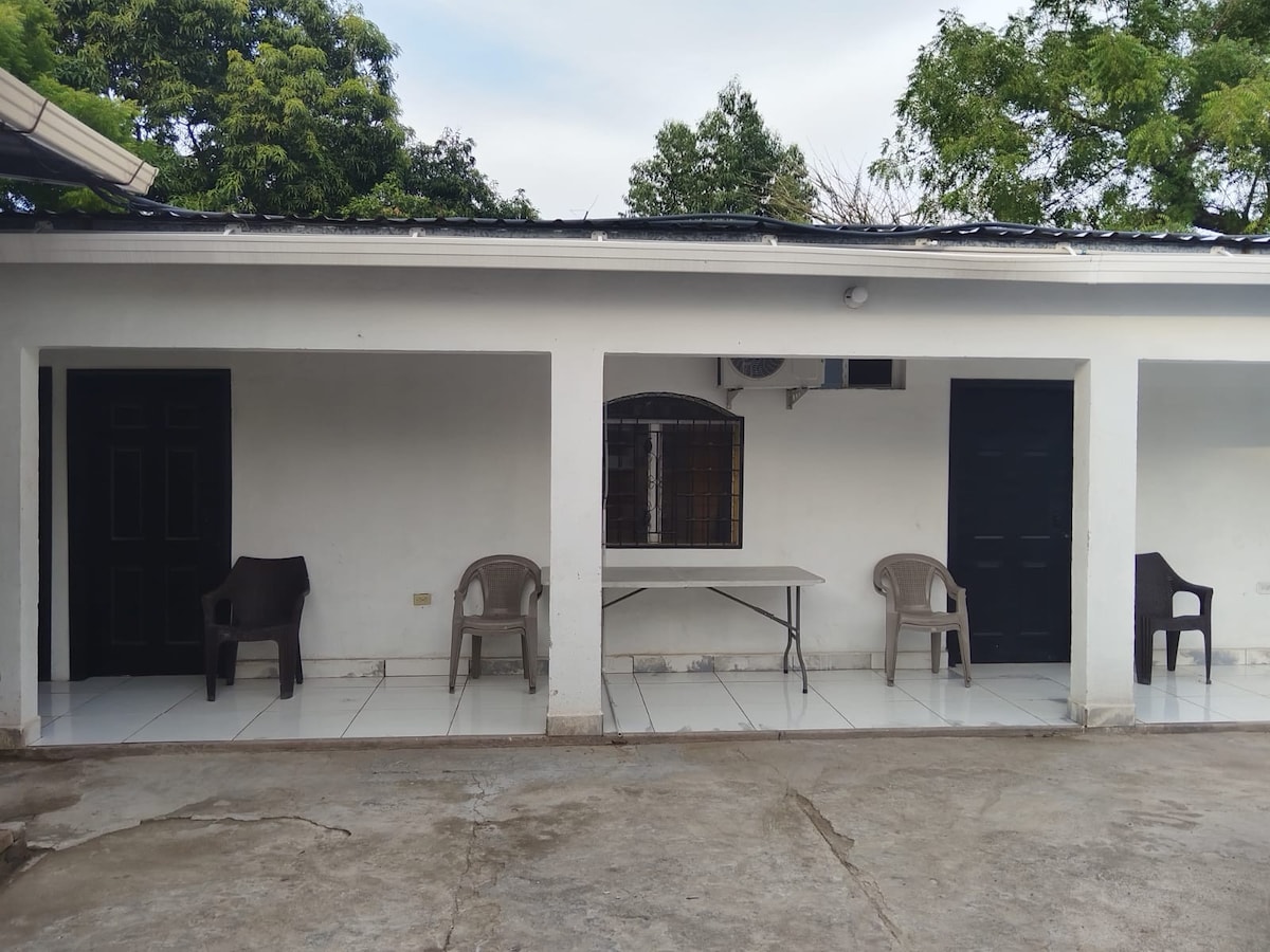 An exterior view of a building features a series of private entrances, each flanked by simple, dark doors. Two light-colored plastic chairs and a table are positioned on the tiled patio, while greenery surrounds the area, enhancing the peaceful environment.