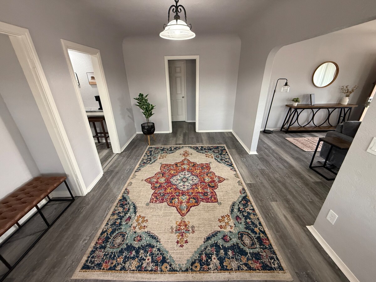 A spacious entryway is adorned with a large, decorative area rug featuring intricate patterns and colors. Light enters through a nearby door, illuminating the neutral walls and wooden flooring. A small seating area is positioned to the left, and a round mirror is visible in the adjoining room.