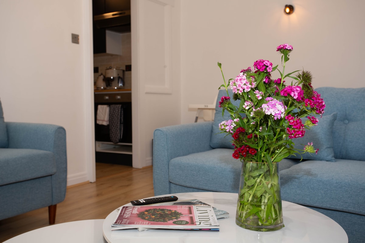 A bright living area is presented, featuring a coffee table topped with a floral arrangement of pink and purple blooms. Two light blue sofas flank the room, while a glimpse of a kitchen area is visible in the background.