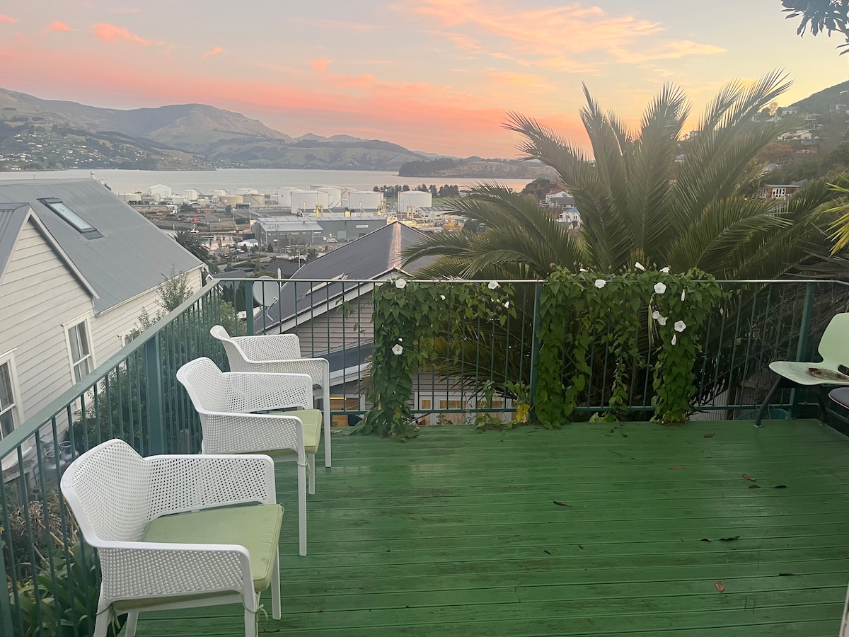 A deck area is showcased, featuring three white chairs adorned with green cushions. Lush greenery climbs along the railing, and views of Lyttelton Harbour and surrounding hills are visible in the background, with soft colors of sunset illuminating the sky.