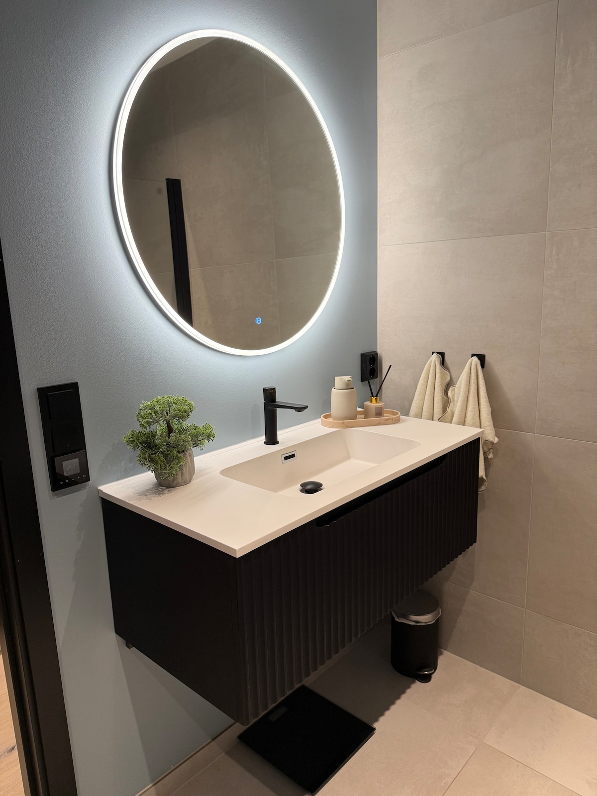 A modern bathroom features a sleek, wall-mounted vanity with a minimalist basin. A round mirror with backlighting is positioned above the sink. Neutral-toned tiles create a clean backdrop, accentuated by a small plant and toiletries on the countertop.