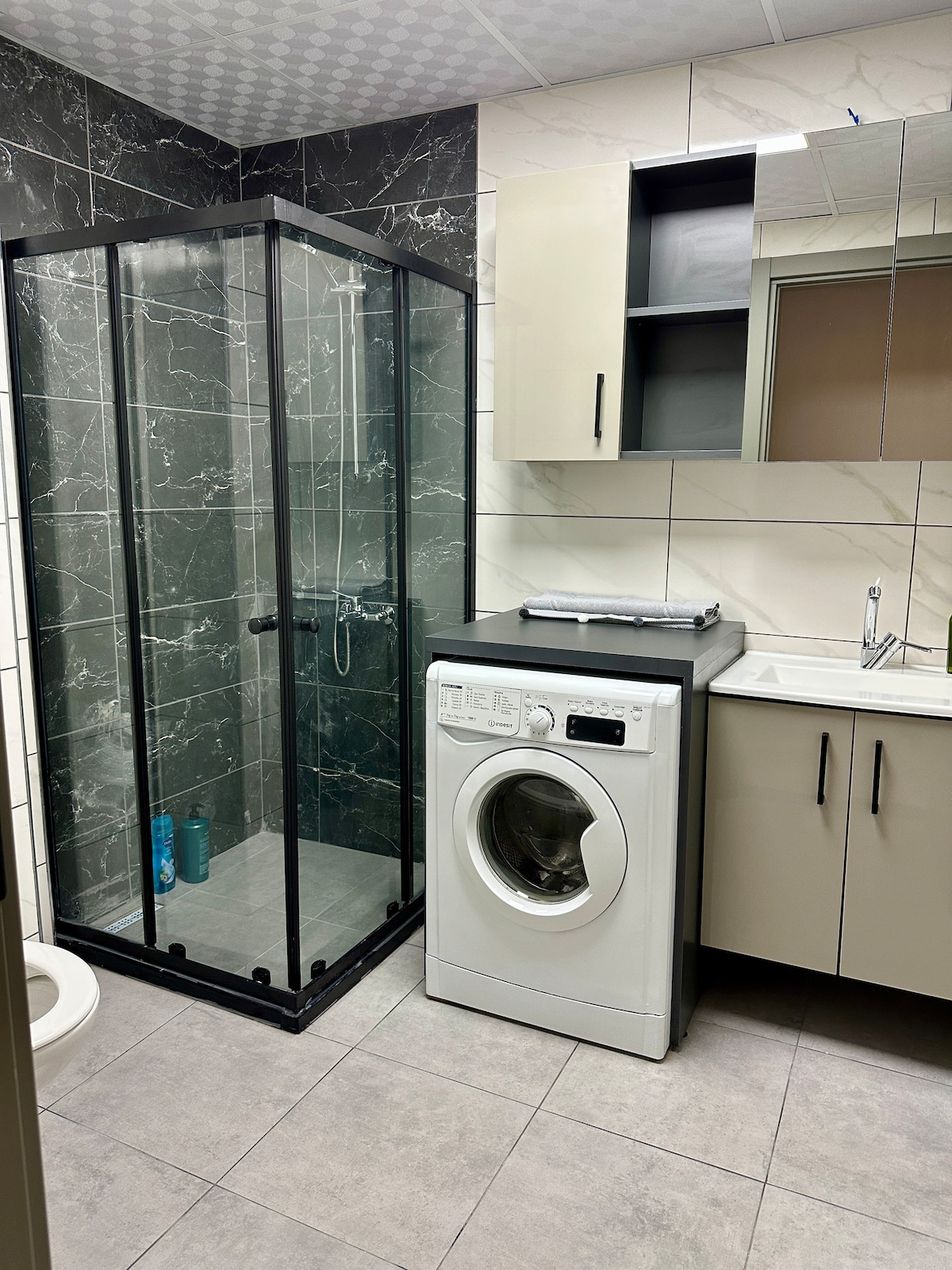 A modern bathroom is presented with a glass shower enclosure, featuring black frames that contrast with the light tiles. A washing machine is positioned next to a sleek vanity with a sink, while wall-mounted cabinets provide additional storage.