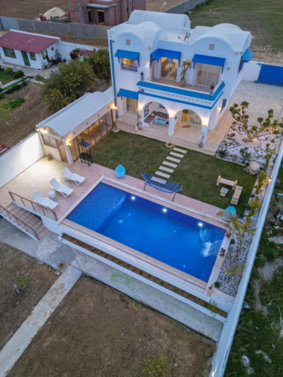 An aerial view of a spacious villa features a shimmering pool surrounded by a sun deck and lounge chairs. A well-maintained garden complements the outdoor area, while a shaded patio space offers seating. The villa showcases architectural elements typical of Arabic style.