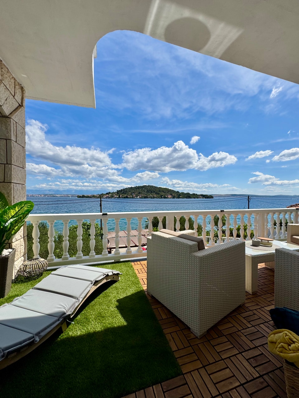 A balcony area is shown, featuring modern outdoor furniture including two chairs and a chaise lounge. The space is complemented by green grass flooring and a view of the ocean, with an island visible in the distance under a partly cloudy blue sky.