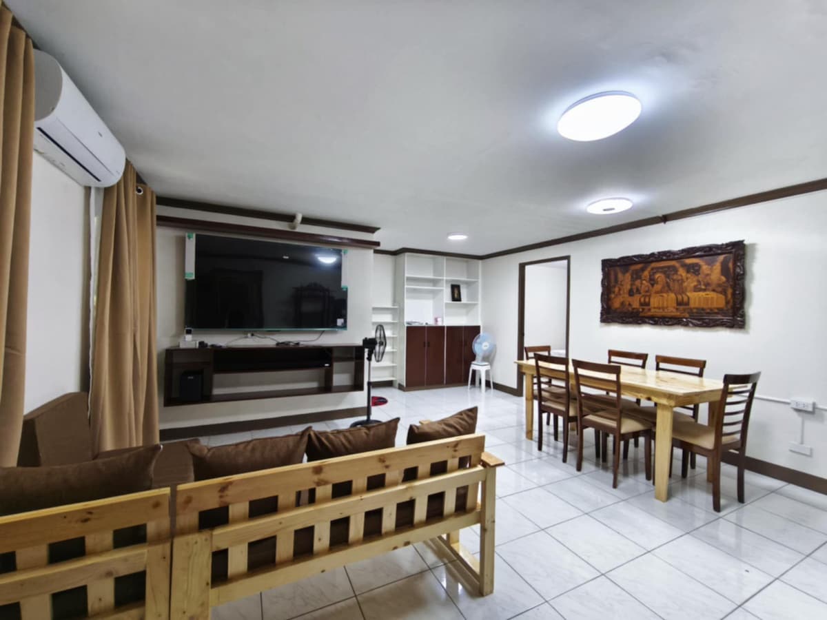 A spacious living area is equipped with a wall-mounted television and wooden dining table surrounded by multiple chairs. Natural light filters through curtains on the window, illuminating the neutral tones of the walls and tiled floor. An air conditioning unit is mounted on the wall.