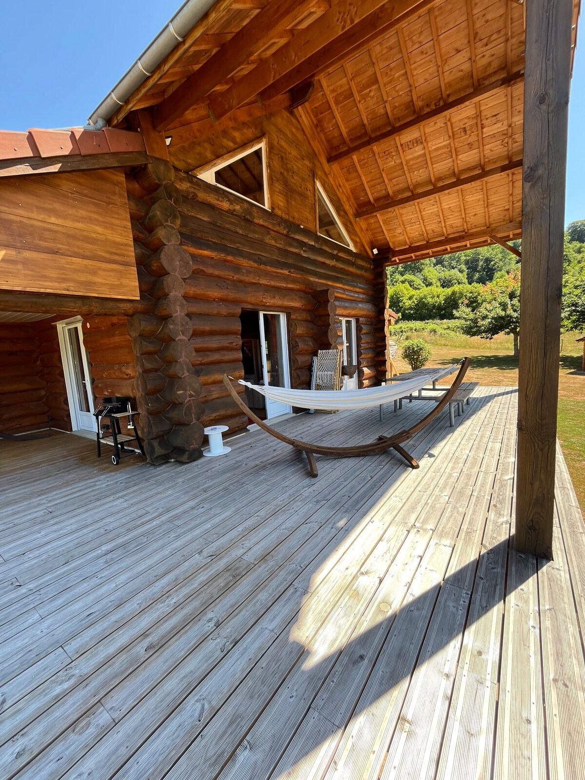 A spacious wooden deck extends from the cabin, featuring a long table and chairs for outdoor dining. A hammock is positioned for relaxation, while large glass doors lead into the interior. Lush greenery is visible in the background, complementing the natural setting.