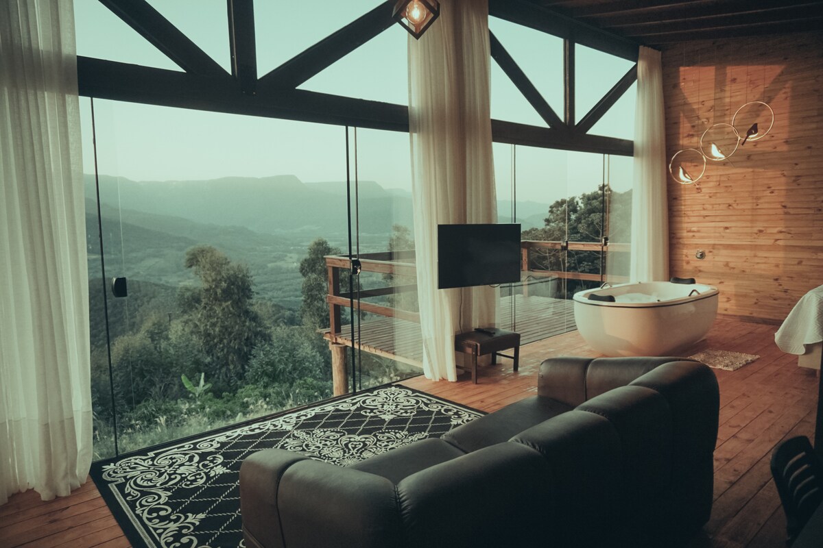 A serene living space is enhanced by large glass windows offering expansive mountain views. A modern sofa sits comfortably on a patterned rug, while a stylish bathtub is positioned near the window. Soft curtains diffuse natural light, creating a peaceful ambiance.