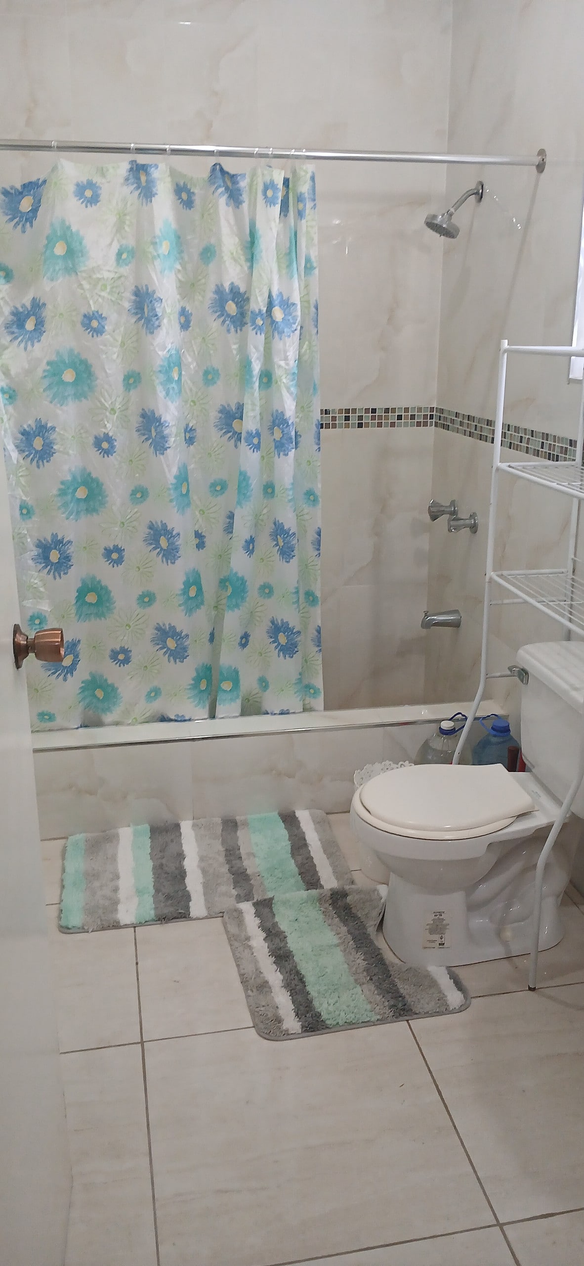 The bathroom features a shower with a colorful floral curtain, a white toilet, and a compact shelving unit for storage. Soft, striped mats are laid on the tiled floor, and small tiles decorate the wall above the shower area.