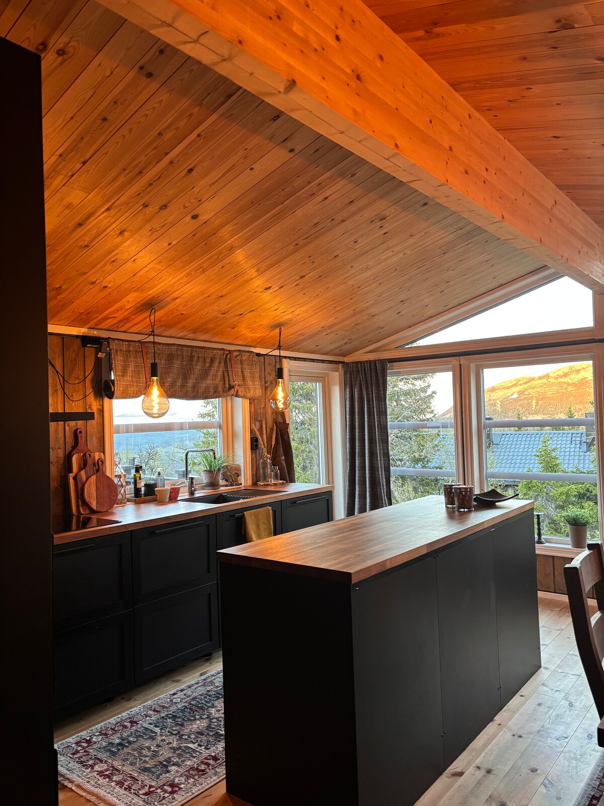 A modern kitchen features a wooden ceiling with a slanted design, enhancing the open feel of the space. Counters are lined with simple decor and appliances, while natural light floods through large windows, offering views of the surrounding landscape.