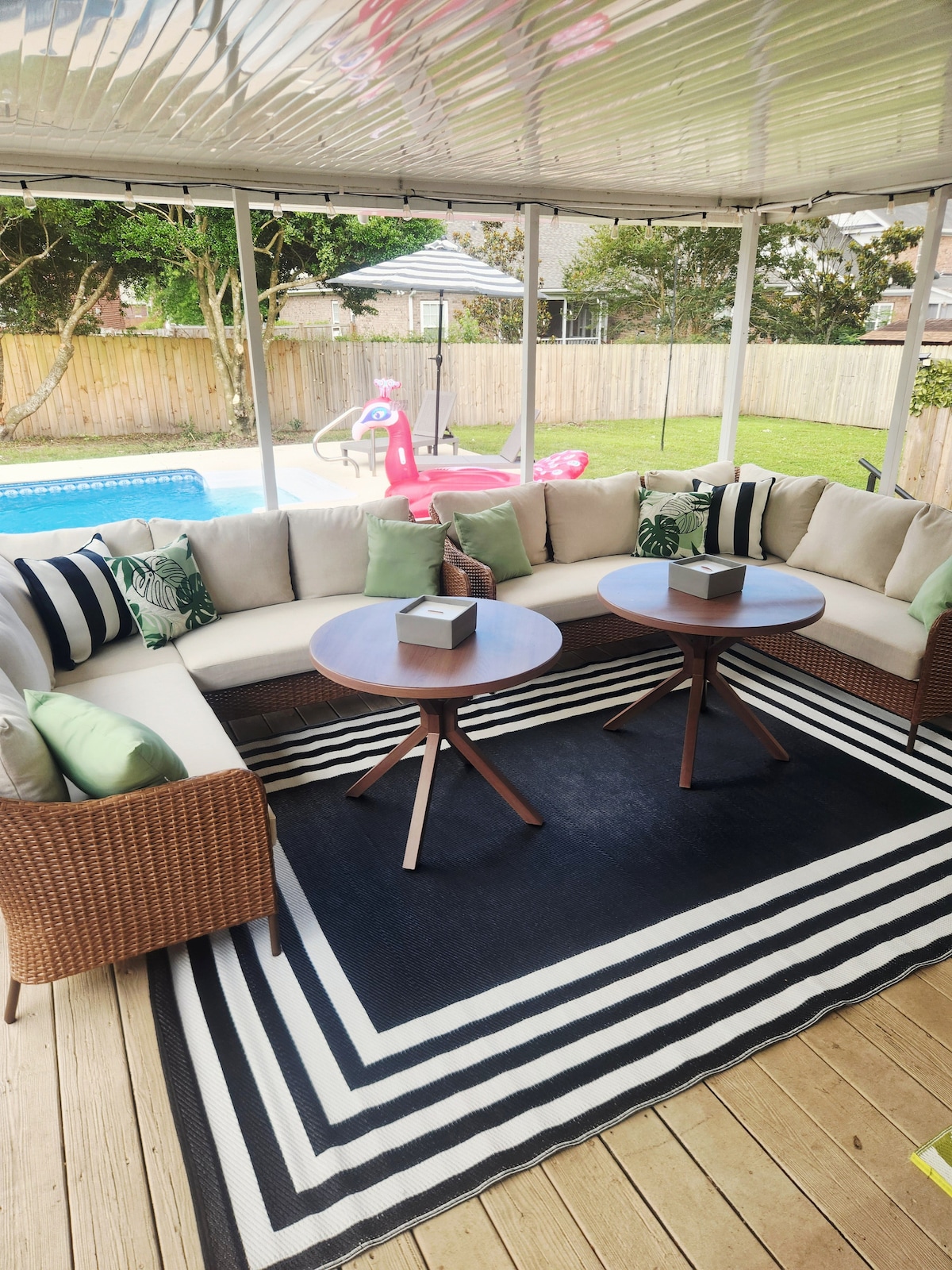 A screened-in porch features a large, circular sectional sofa with green and black accent pillows. Two circular tables are placed on a striped rug, adding a cozy touch. The background shows a private pool area bordered by grass and a wooden fence.