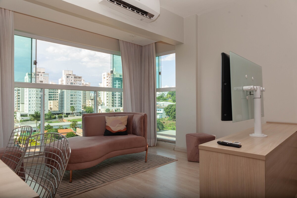 A light-filled living area features a large window with views of the cityscape. A comfortable sofa is positioned in front of a modern TV mounted on the wall. A wooden desk holds a sleek monitor and remote, contributing to a functional workspace.