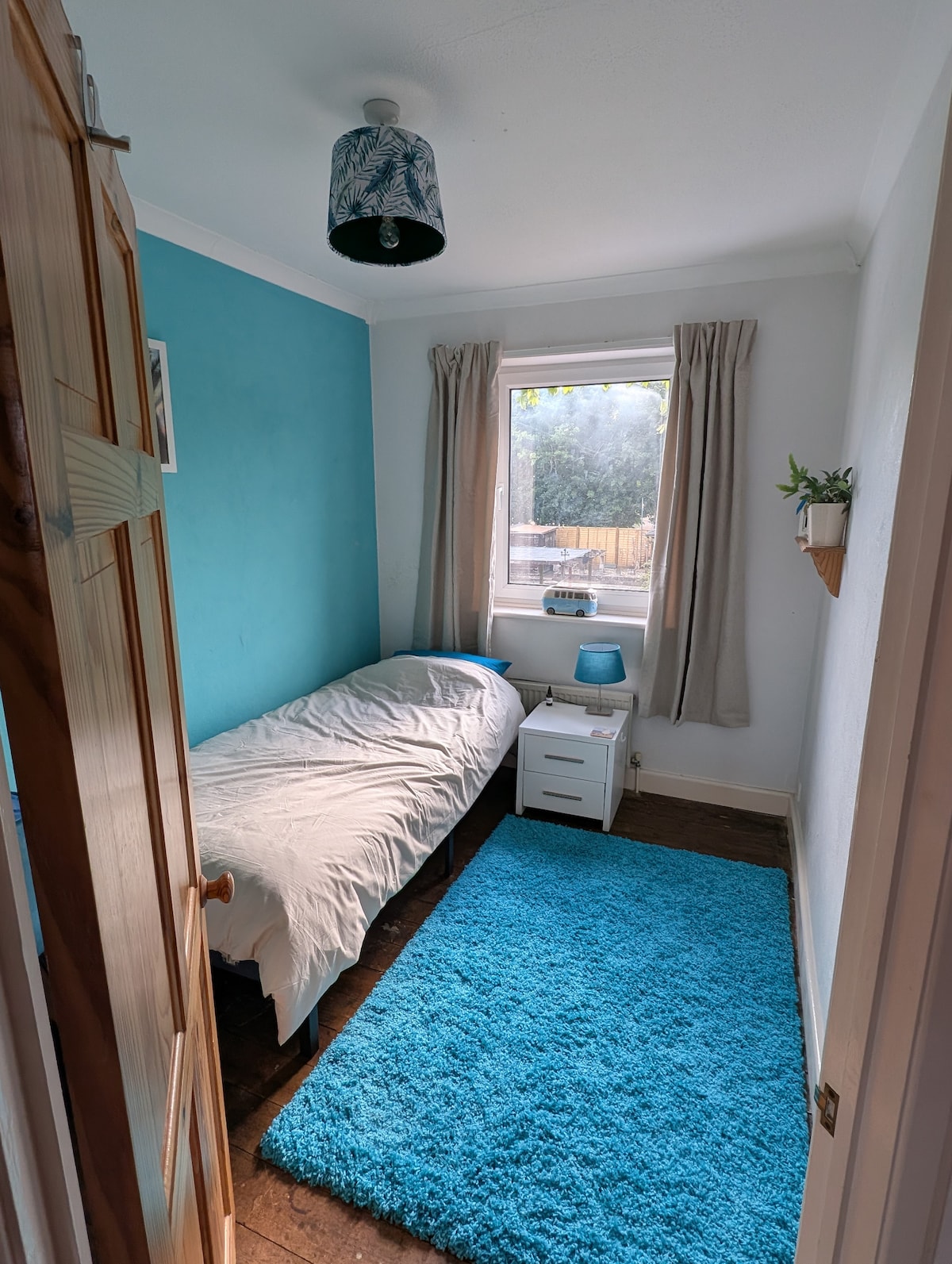 A cozy bedroom features a single bed dressed in light bedding. A large window allows natural light to enter, offering a view of the outdoors. A small nightstand with a lamp stands beside the bed, and a vibrant blue carpet covers the floor.