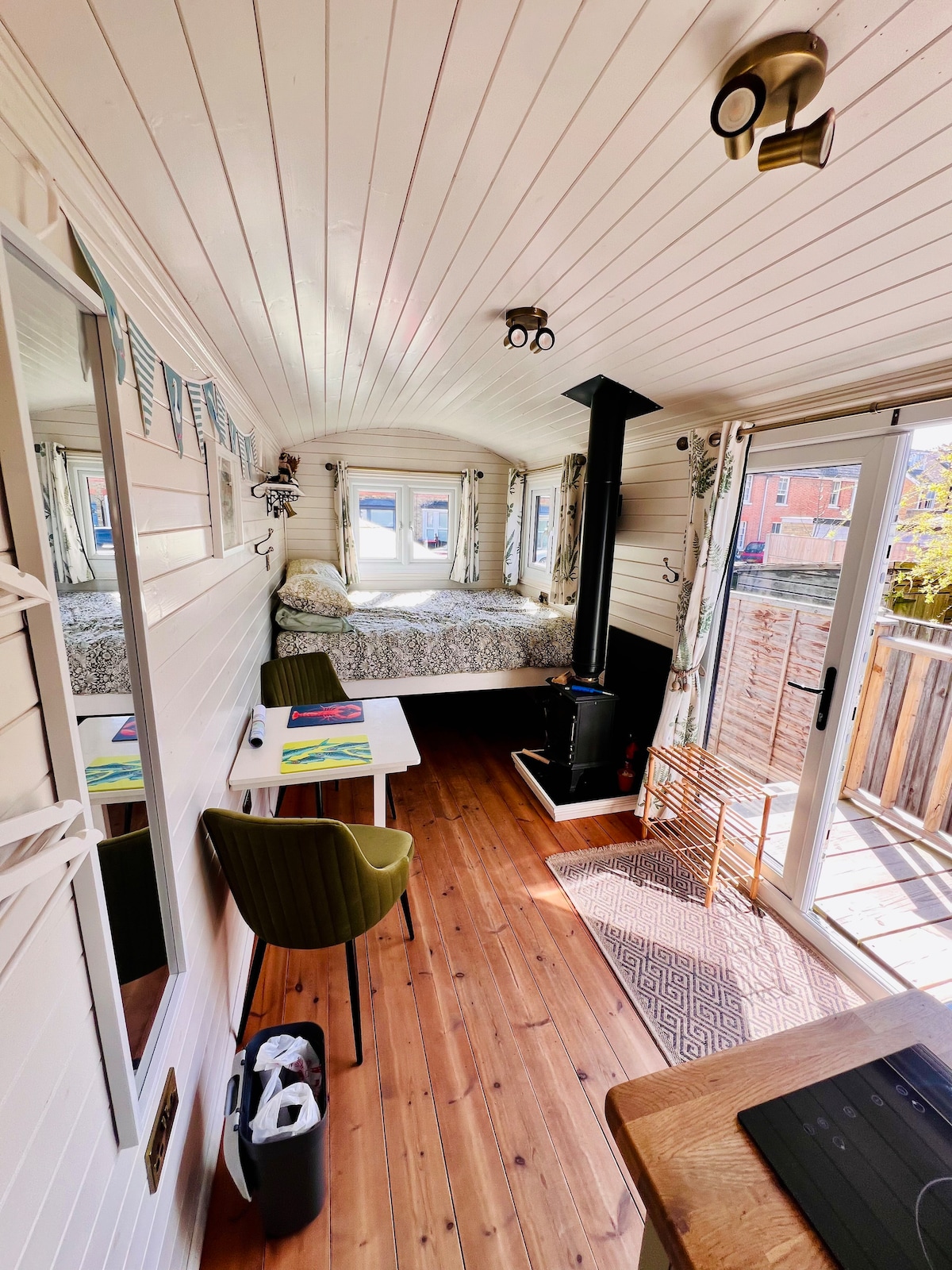 The interior of the shepherd's hut showcases a cozy sleeping area with a spacious bed, accented by decorative pillows. A wood-burning stove provides warmth, while a small dining table for two sits next to a sliding glass door, leading to the private outdoor space.