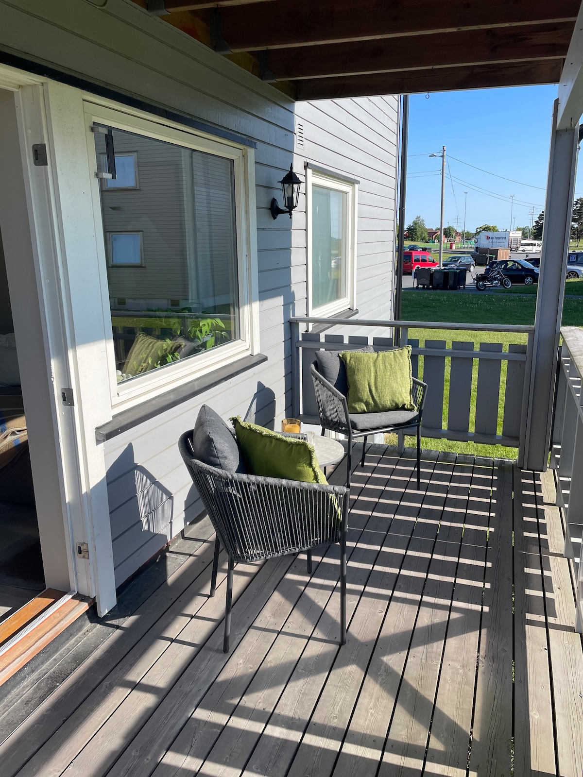 A small outdoor balcony is furnished with two black chairs, each complemented by green cushions. Natural light filters through the sliding glass door, highlighting the wooden floor. A view of the surrounding area is visible, featuring greenery and distant structures.