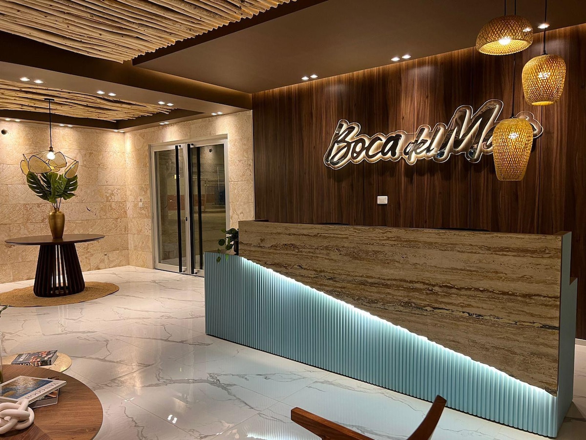 A modern reception area is presented with a sleek front desk featuring a textured surface. Warm lighting is provided by pendant fixtures above, while natural elements, such as a plant, are incorporated into the design. Large elevators are positioned to the left, enhancing accessibility.