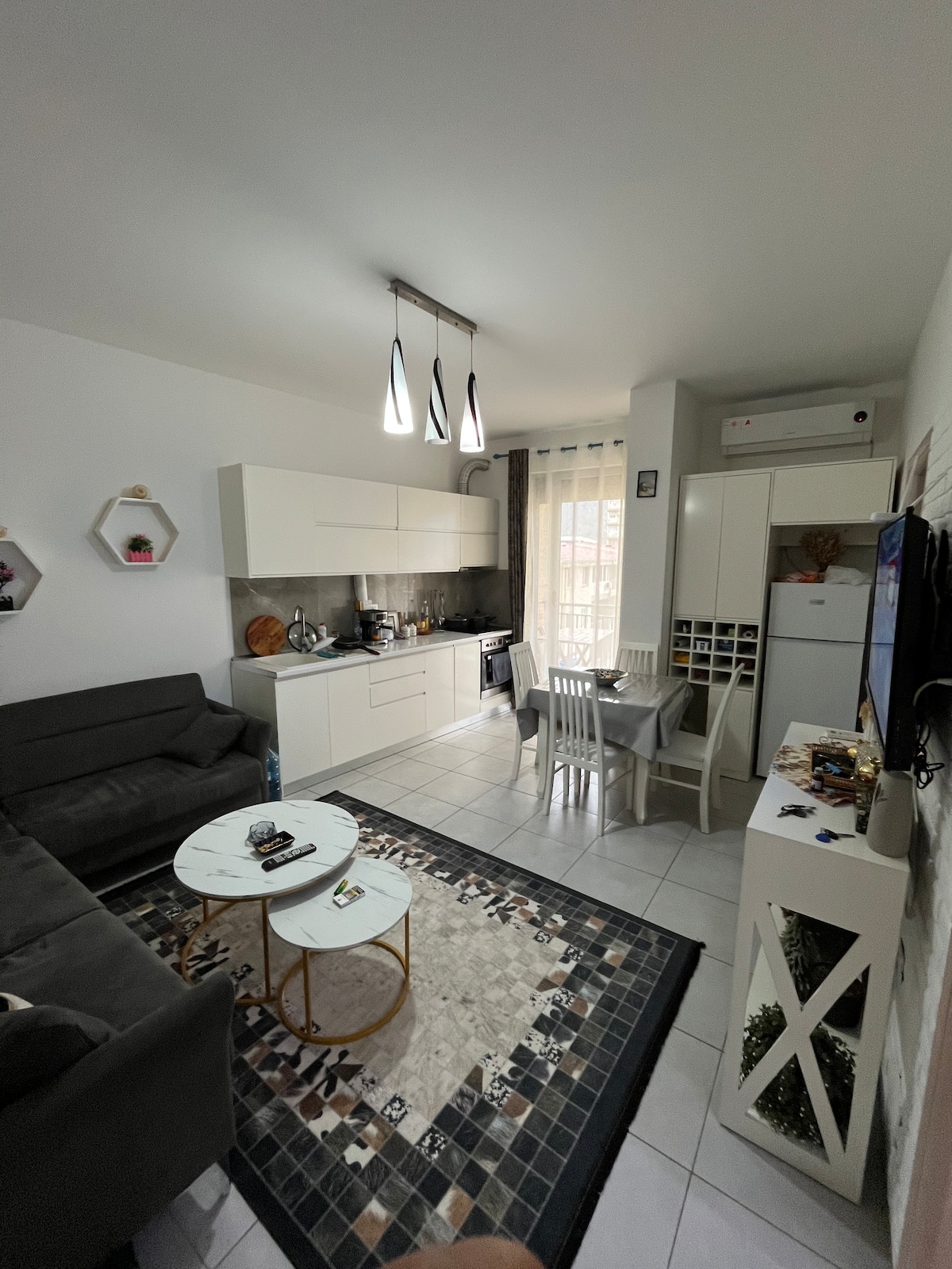 A compact living area features a dark gray sofa and a small dining table beside a kitchen with modern cabinetry. Soft light is provided by pendant fixtures above, and a patterned rug adds texture to the tiled floor. A window allows natural light to enter.