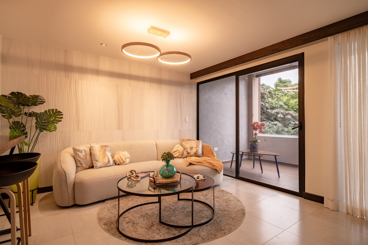 The living area features a modern sectional sofa adorned with decorative pillows, a circular coffee table, and a soft area rug. Large glass doors open to a terrace, inviting natural light and green outdoor views, complemented by gentle lighting fixtures overhead.