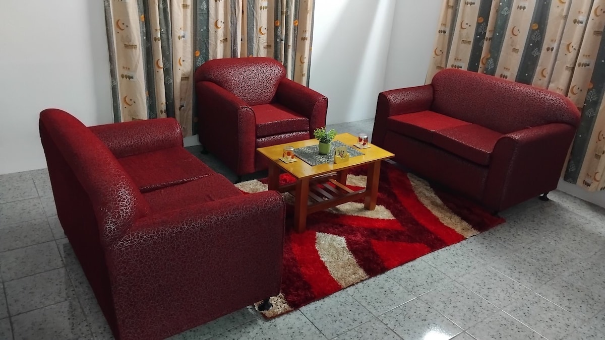 A cozy seating area features two red armchairs and a matching sofa, arranged around a wooden coffee table. A vibrant red and white patterned rug enhances the space. Soft curtains with a subtle design frame the windows, allowing natural light to illuminate the room.