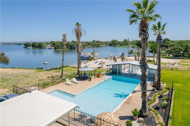 A spacious outdoor area features a shimmering pool bordered by lounge chairs and palm trees. The tranquil waters of Lake LBJ are visible in the background, providing a serene backdrop. A grassy lawn extends towards the water, enhancing the inviting atmosphere of the surroundings.