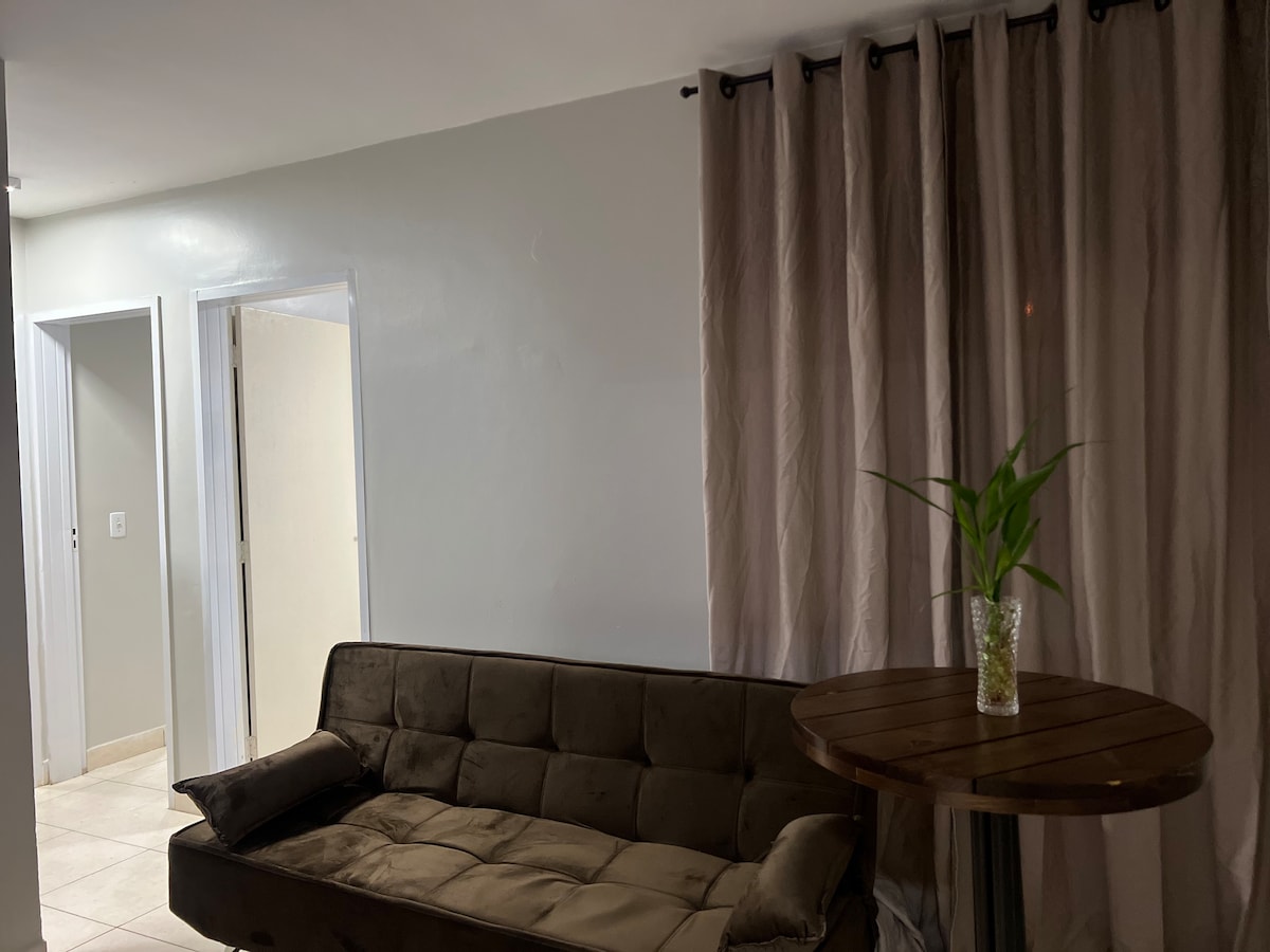 A cozy seating area is presented, featuring a dark sofa positioned near a small wooden bistro table. Light is softly diffused through sheer curtains, enhancing the quiet ambiance. The room is complemented by a decorative plant on the table, contributing to the overall inviting feel.