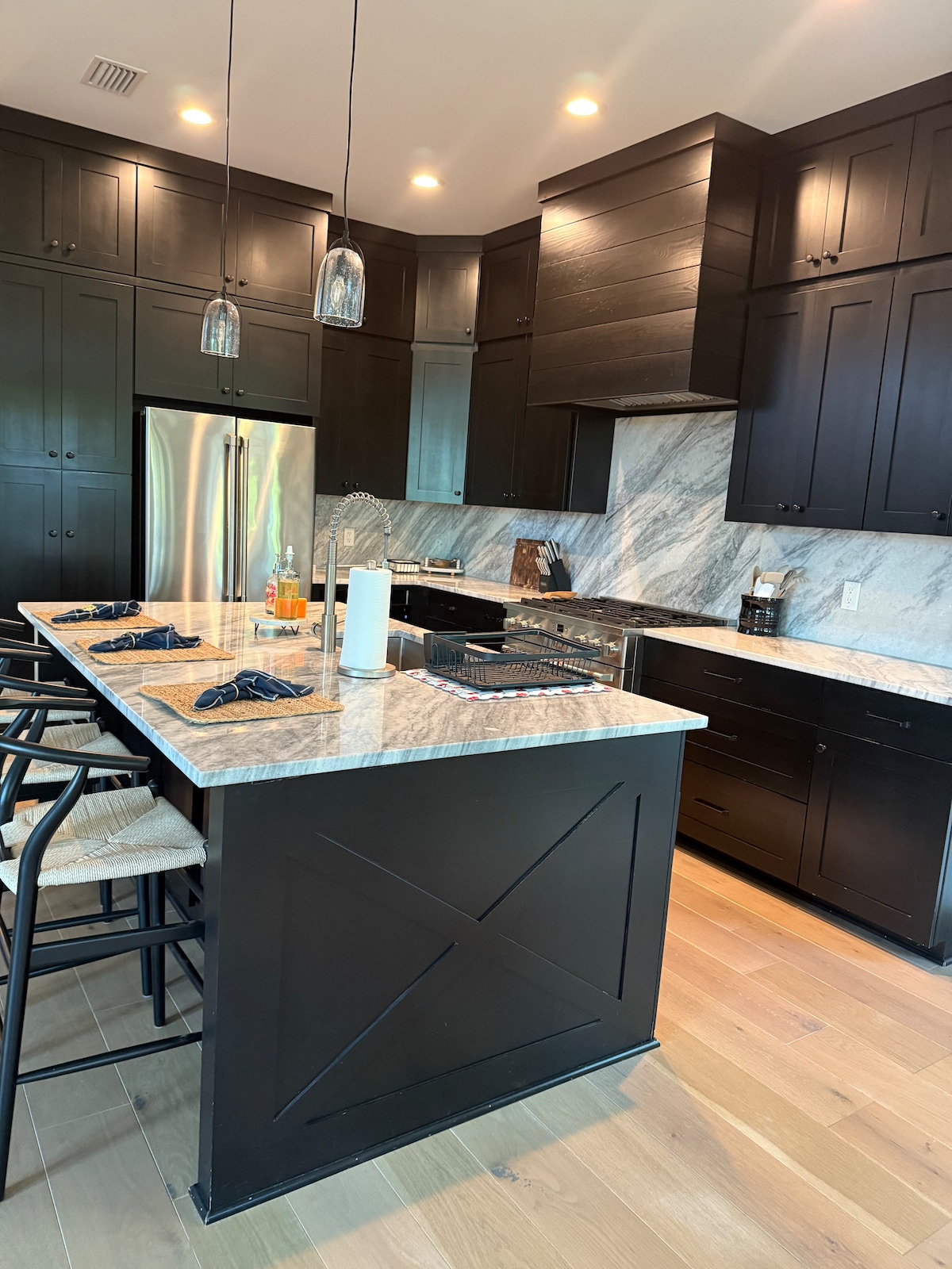 A modern kitchen features dark cabinetry paired with a light marble backsplash. An island with seating is highlighted, and pendant lights offer illumination. Stainless steel appliances are included, with a silver refrigerator and a built-in oven. The wooden flooring adds warmth to the space.