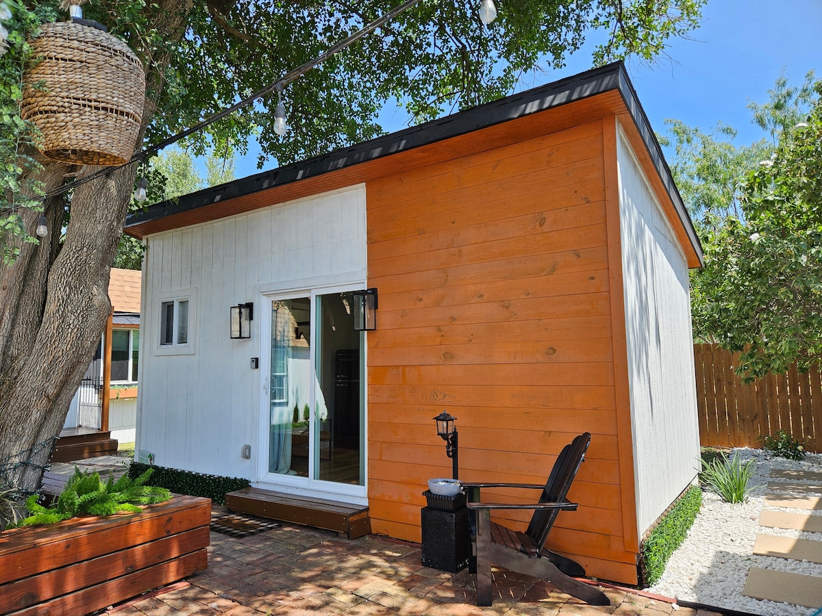 A modern guest house presents a combination of white and wooden siding, contrasting with the lush greenery surrounding it. A spacious glass door opens onto a small patio area, while natural light highlights the inviting exterior. Decorative elements include a lantern and an outdoor chair.