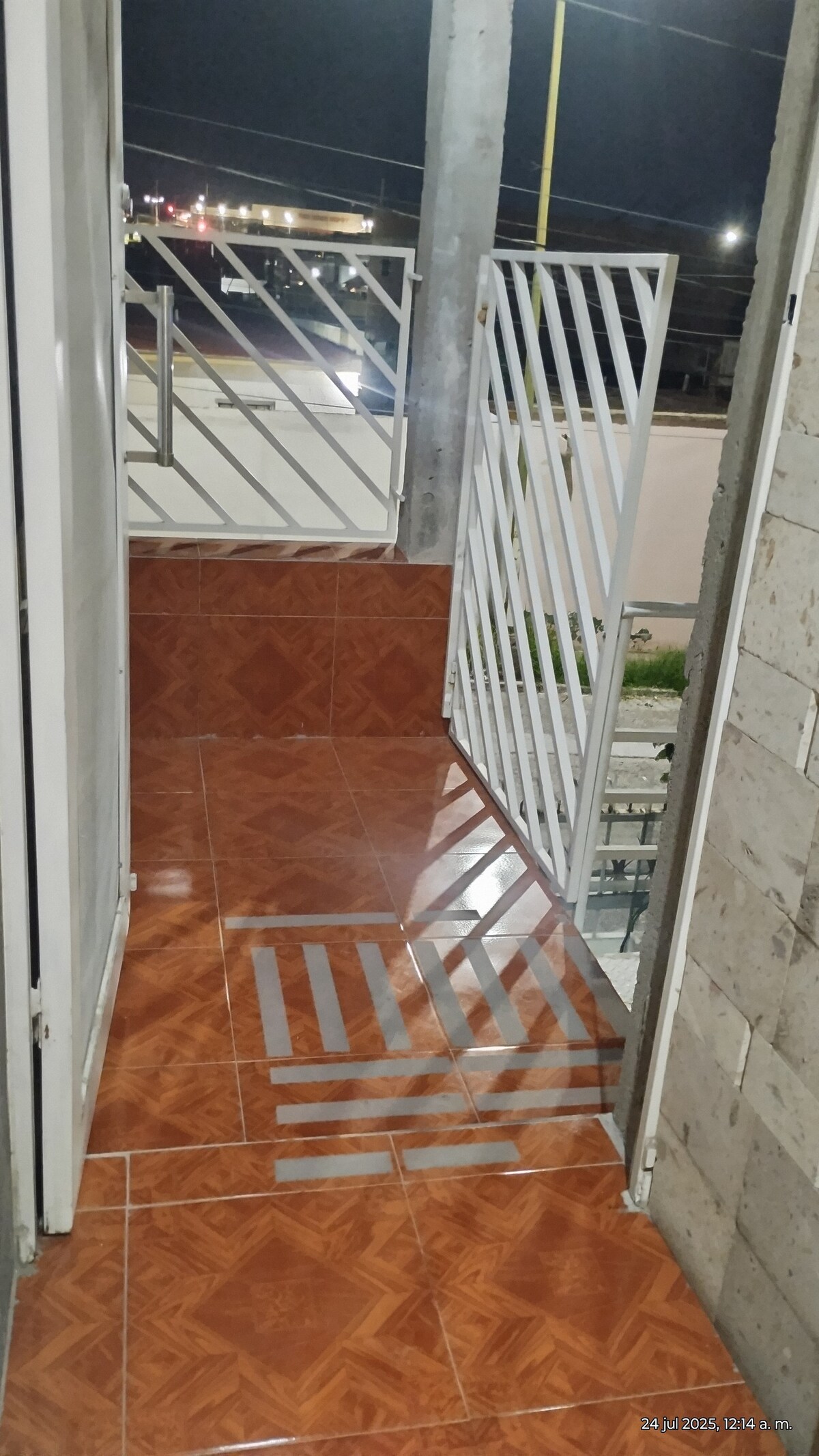 A small terrace is shown, featuring tiled flooring with a shiny finish. A railing is visible, providing safety, while the surrounding area is illuminated by nearby lights. The entrance is framed by two doors that lead to the interior space.