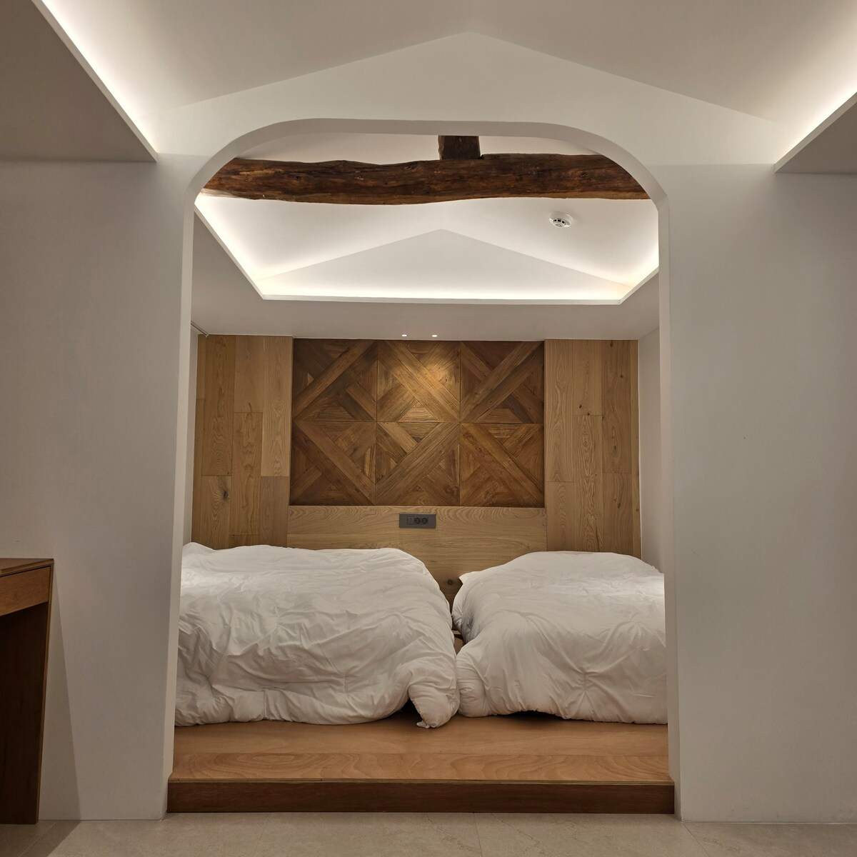 A sleeping area features two spacious beds with softly draped white bedding, positioned beneath a wooden beam. The wall behind is adorned with a geometric wooden panel, adding warmth to the modern design. Subtle lighting along the ceiling provides a gentle glow.