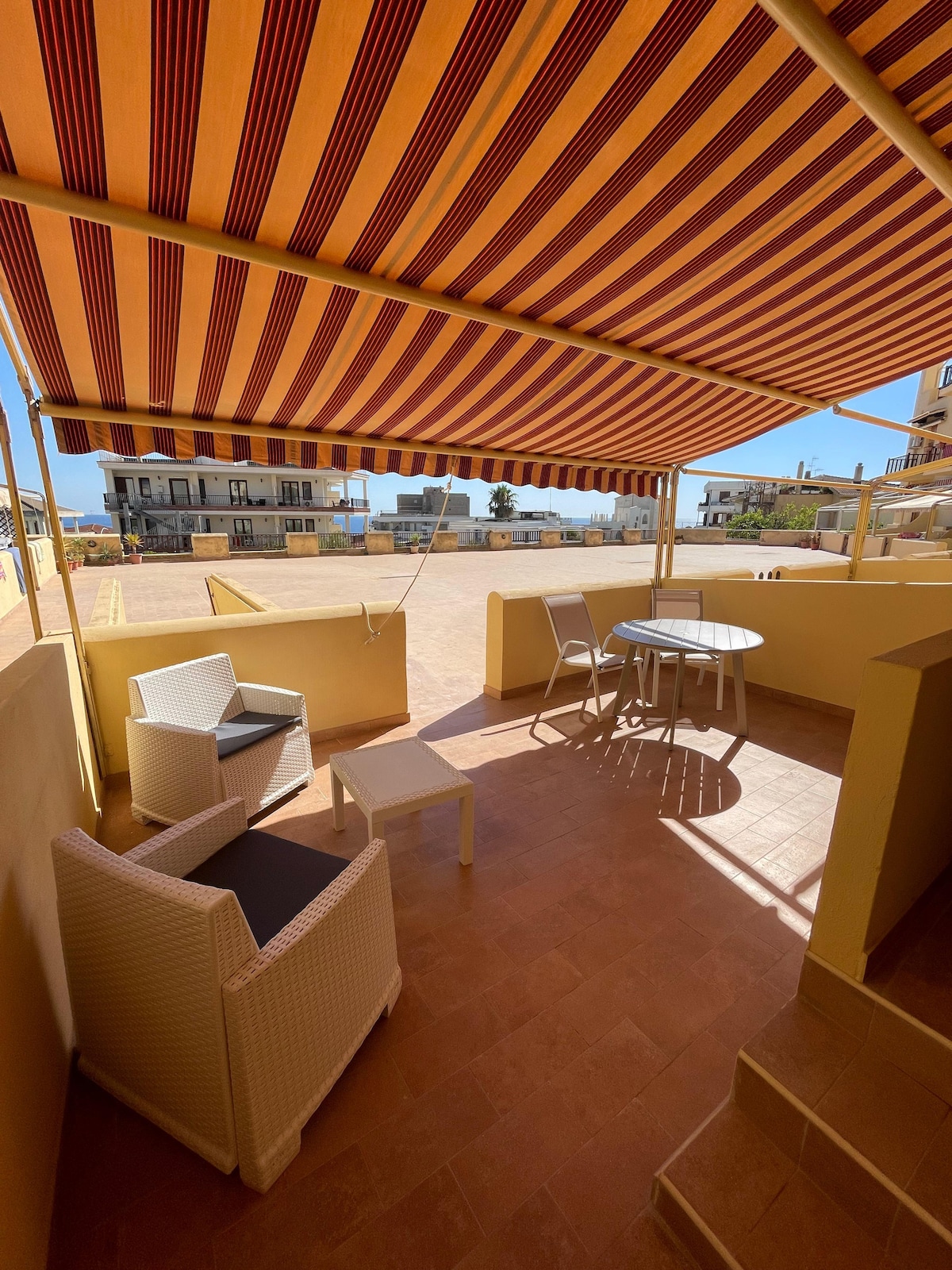The outdoor terrace features a striped awning providing shade, with two white lounge chairs and a small round table. The tiled floor reflects the sunlight, with a clear view of the surrounding area, creating a private space for relaxation.