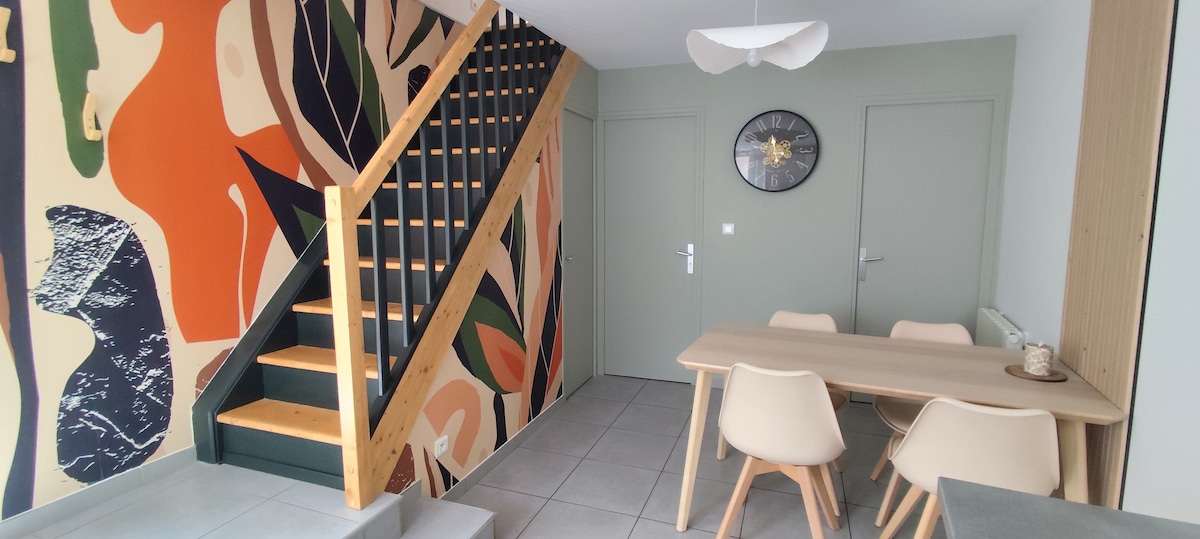 A bright entryway features a staircase with wooden steps and a contemporary metal railing. The walls are adorned with a vibrant mural of abstract shapes. A light wood dining table surrounded by four light-colored chairs is positioned on tiled flooring, complemented by a wall clock and a minimalist light fixture above.