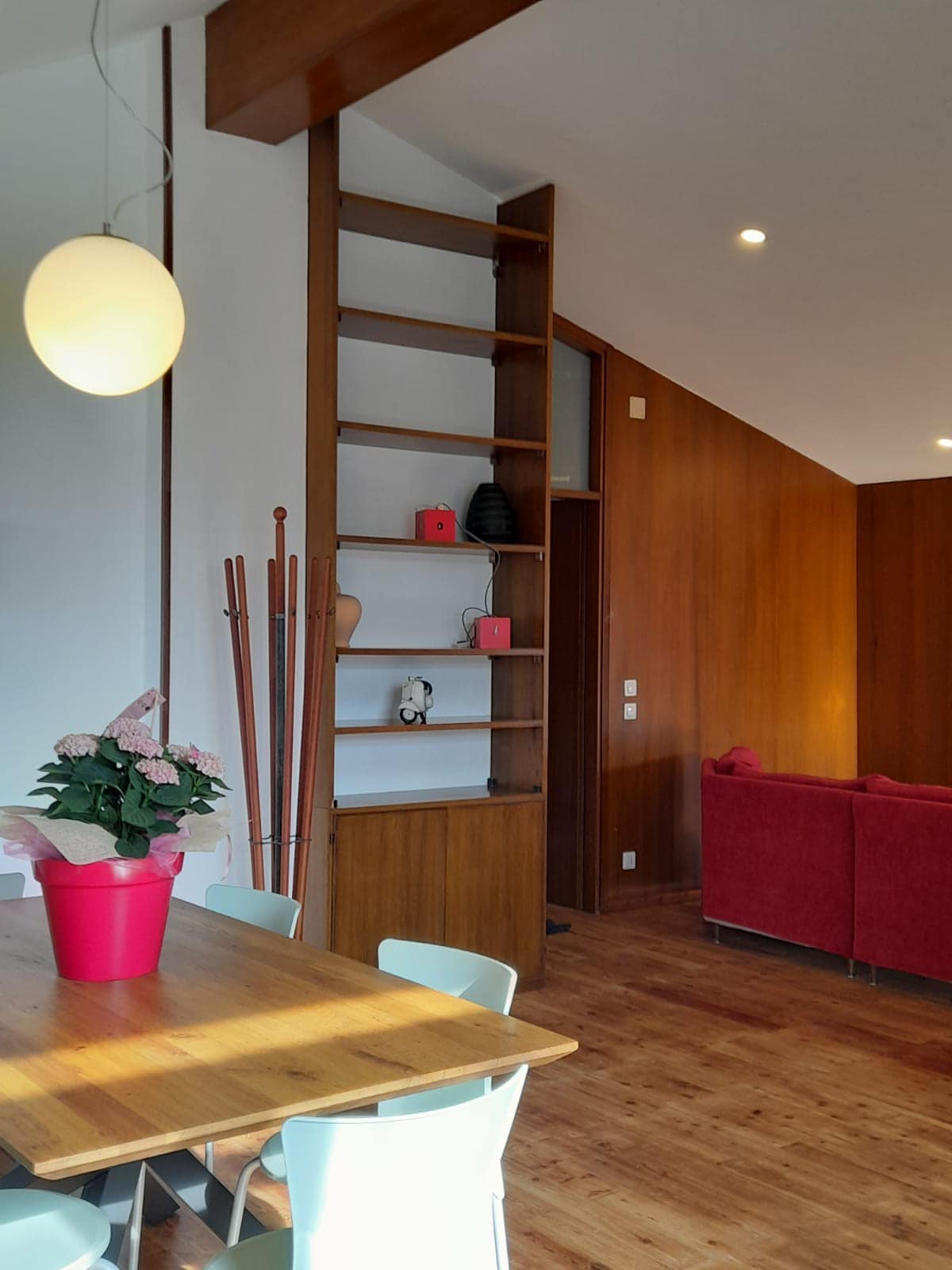 A wooden bookshelf stands against a warm-toned wall, displaying decorative items. A dining table with light-colored chairs occupies the space. A pink plant is positioned on the table, and a cozy red sofa is visible in the background.