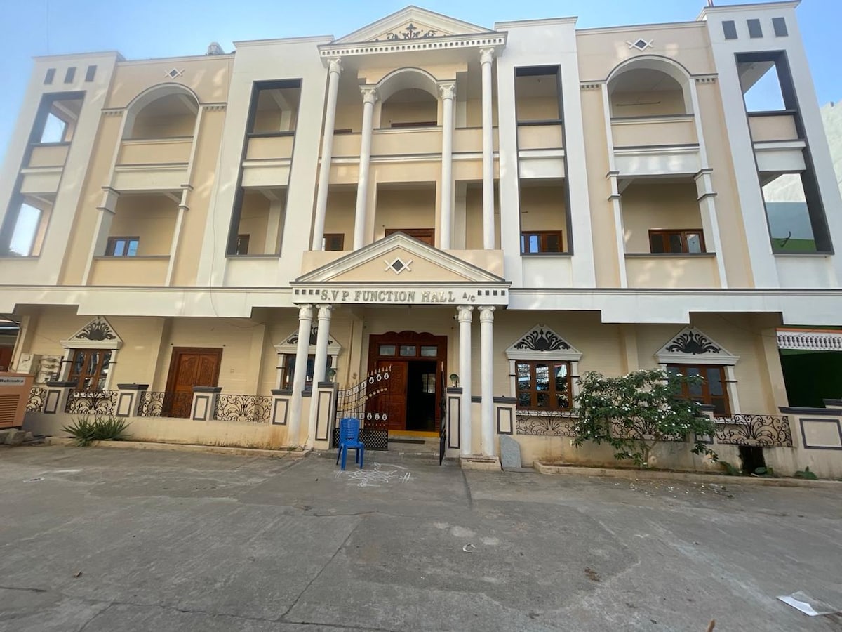 The exterior of a multi-story function hall presents a classic design, characterized by arched doorways and columns. Decorative elements frame the entrance, and greenery is visible along the sides. The building's façade features neutral tones, creating an approachable ambiance.
