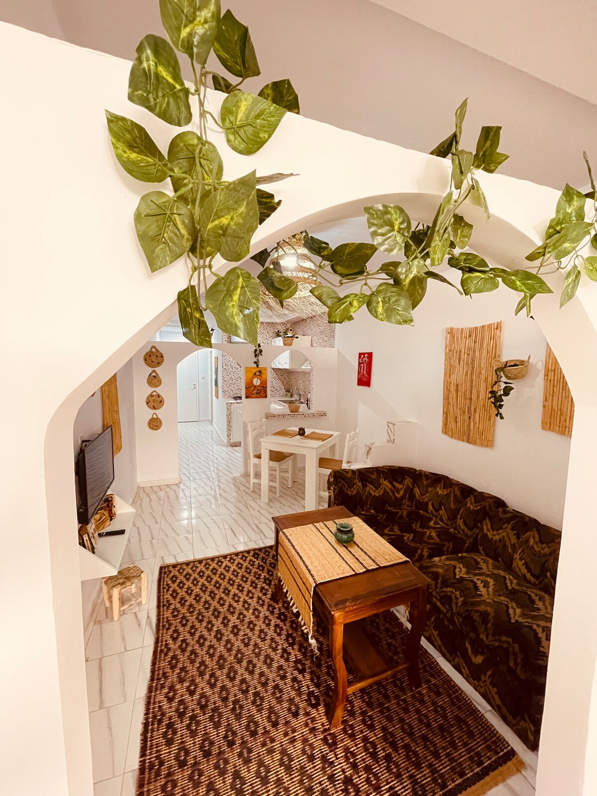 An inviting interior showcases an archway leading into a cozy living area. A patterned rug covers the floor, complemented by an intricately designed brown sofa. A wooden coffee table sits at the center, with potted plants hanging above, enhancing the room's welcoming atmosphere.