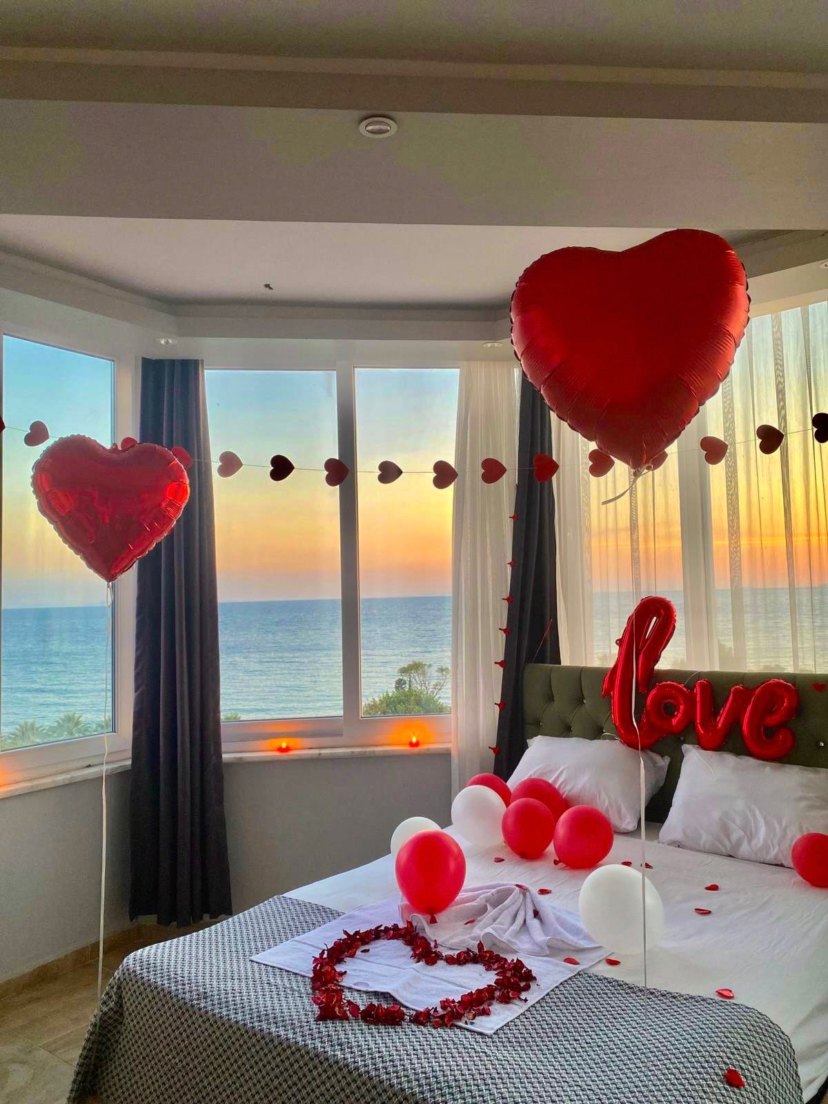 A well-lit bedroom offers stunning ocean views through large windows adorned with sheer curtains. Heart-shaped balloons and red decor create a festive atmosphere. A spacious bed features decorative pillows and a floral arrangement. Candles are placed on the bedside table, enhancing the tranquil setting.