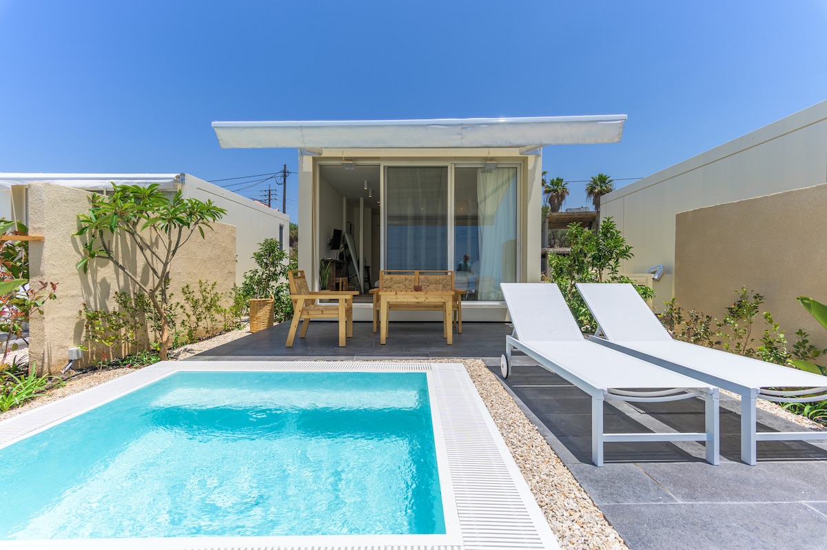 A private plunge pool is framed by a stone patio and surrounded by lush greenery. Two lounge chairs are positioned near the pool, with a small wooden table set adjacent to the entrance of a modern unit featuring large glass doors that invite natural light.