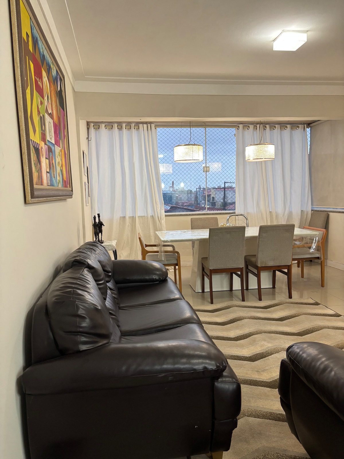 A spacious living and dining area features a dark leather sofa and a dining table with six chairs. Large windows provide natural light, framed by sheer white curtains. Art decorates the wall, adding a colorful touch to the room.