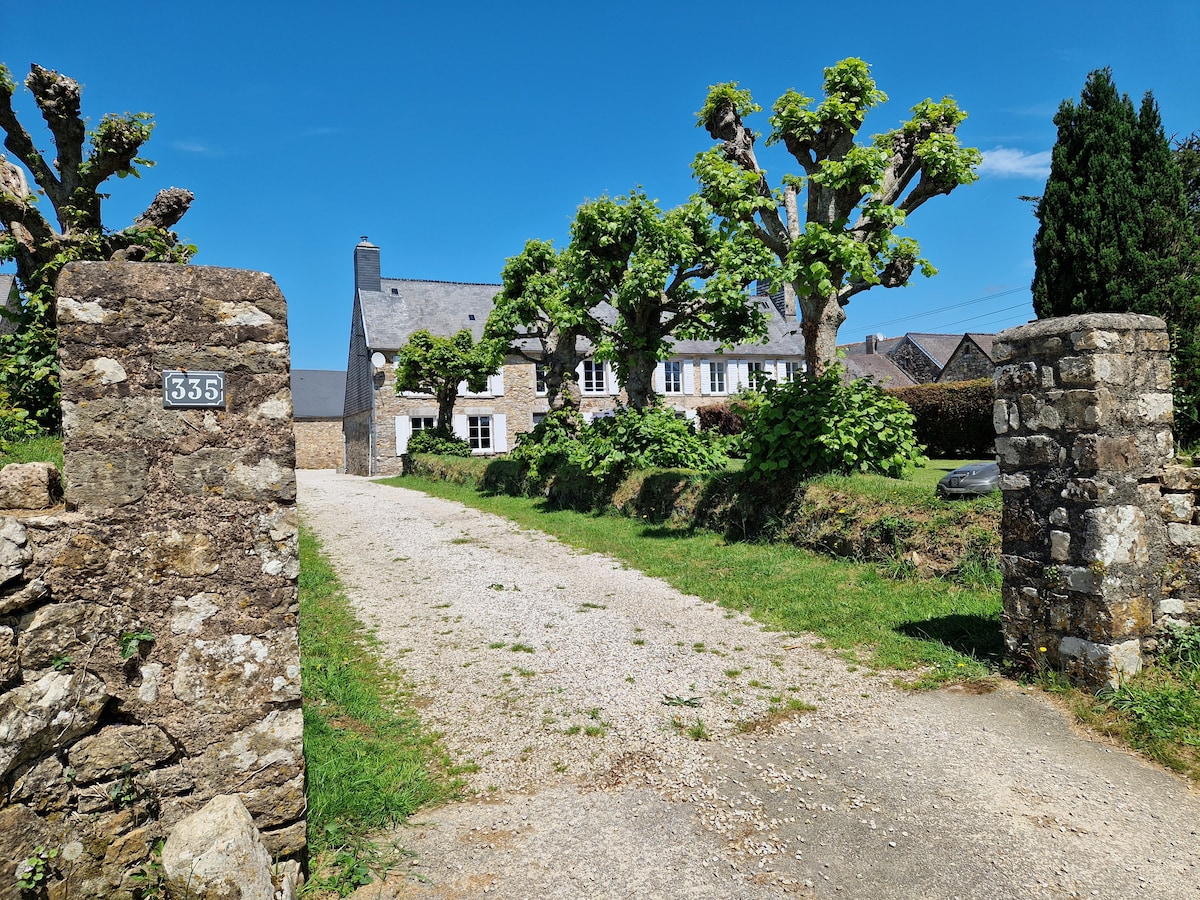 A gravel driveway leads to a charming stone house framed by lush green trees. The entrance is marked by weathered stone gates, featuring the number 335. The clear blue sky provides a bright backdrop for the inviting rural setting.
