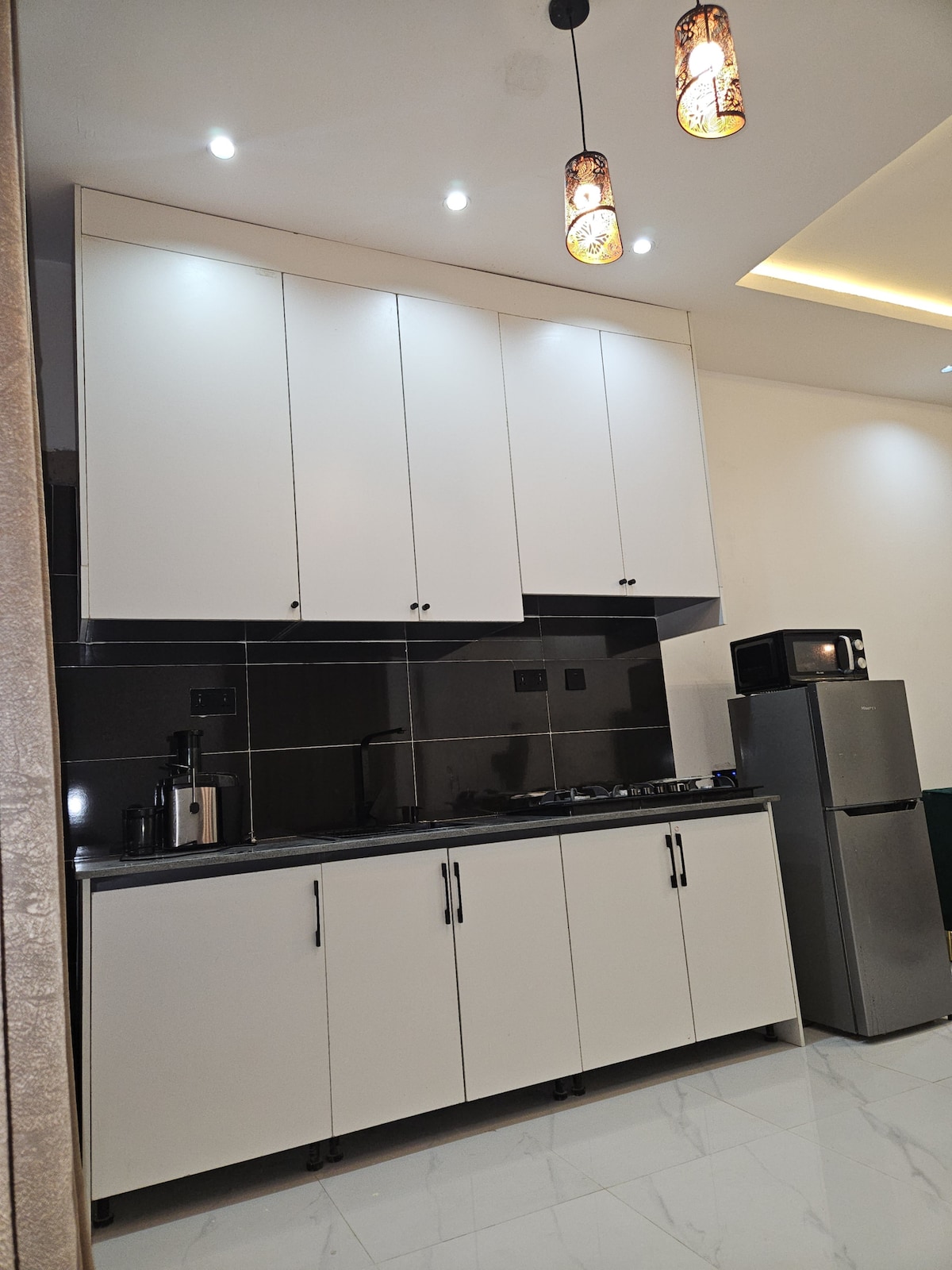 The kitchen features modern cabinetry with ample storage, complemented by a sleek black countertop. Essential appliances, including a refrigerator and microwave, are visible, along with a stovetop. Soft lighting from pendant fixtures highlights the clean and functional design.