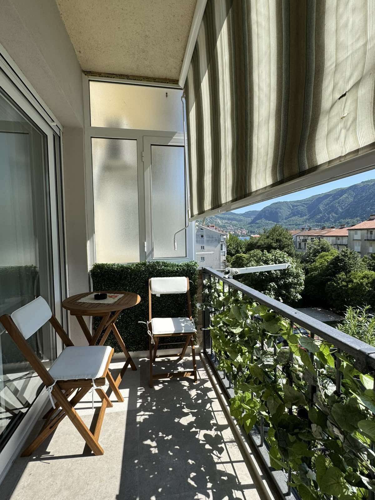 A small terrace is depicted, featuring two foldable chairs and a round table. Green foliage lines the railing, enhancing the space. Sunlight filters through a striped awning, casting gentle shadows on the tiled floor. Scenic views of mountains and buildings are visible in the background.