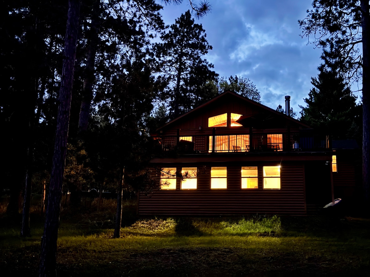 A two-story cabin is set among pine trees, with warm light glowing from large windows. The wooden exterior is accented by a sloped roof, and a deck is visible on the upper level, providing an inviting presence against the evening sky.