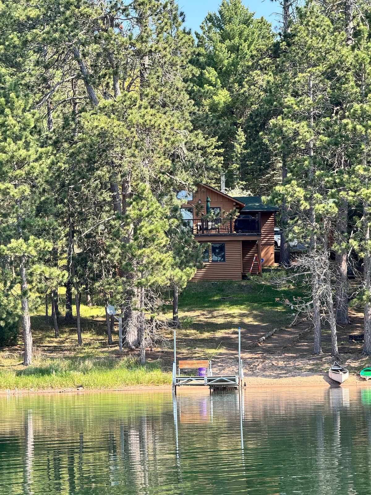 A lakeside log cabin is nestled among tall trees, providing a serene woodland backdrop. The cabin features a deck and large windows offering views of the water. A dock with a small seating area is visible in front, along with canoes resting on the sandy shore.