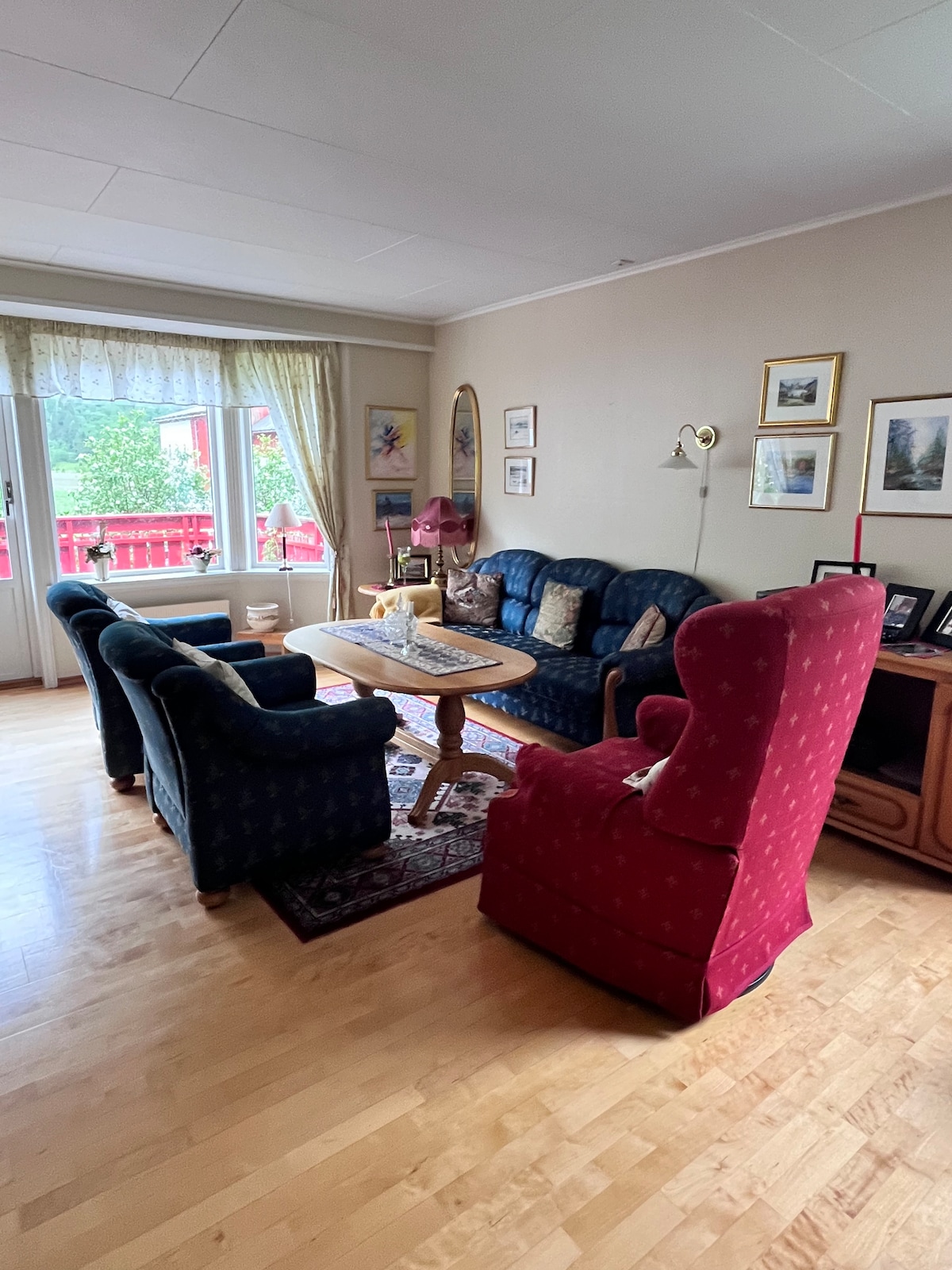 A comfortable living area is showcased with a wooden table surrounded by a mix of seating options, including a red wingback chair and two blue sofas. Natural light floods in through large windows adorned with light curtains, illuminating the warm wooden floor.