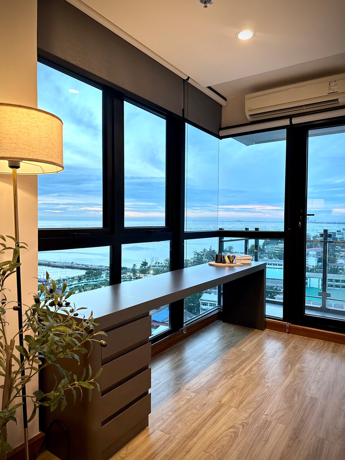 A well-lit workspace is positioned near large windows, providing views of the sea and cityscape. The desk features multiple drawers, while indoor plants add a touch of greenery, enhancing the inviting atmosphere. The flooring is composed of light-colored wood, contributing to a warm ambiance.