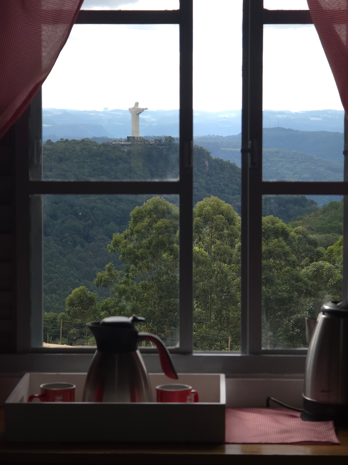 A kitchen window offers a view of the Christ the Redeemer statue in the distance, framed by green mountains. The window features white frames and light curtains, while a tray with a kettle and cups is positioned on the countertop beneath.